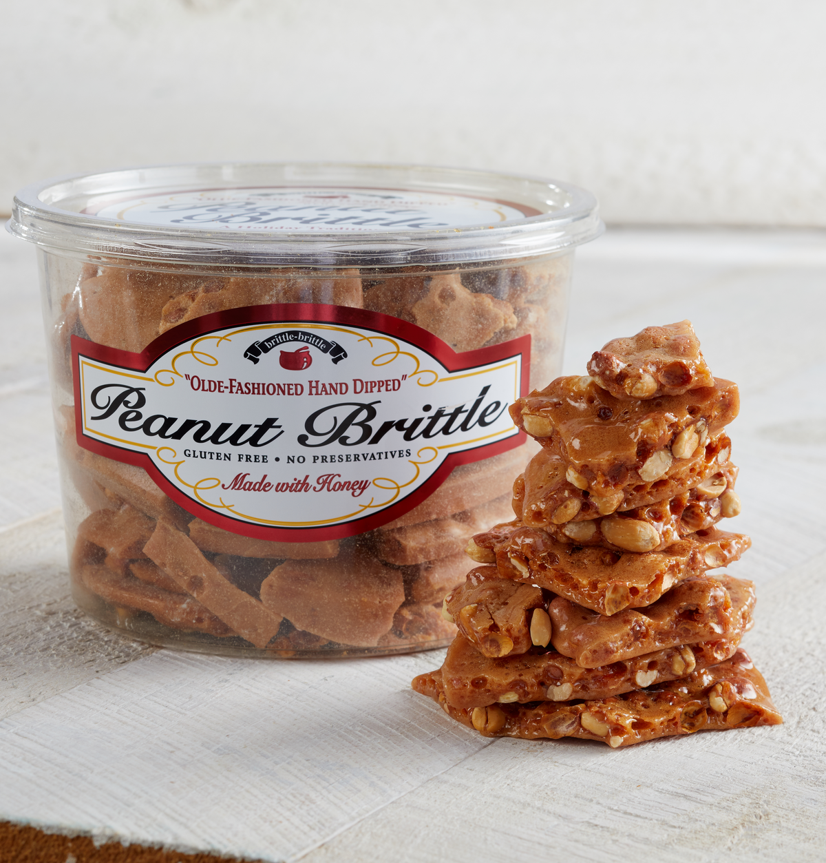 A clear plastic tub of Peanut Brittle with a white, red, and black label, shown next to a tall stack of square peanut brittle pieces on a white textured surface.