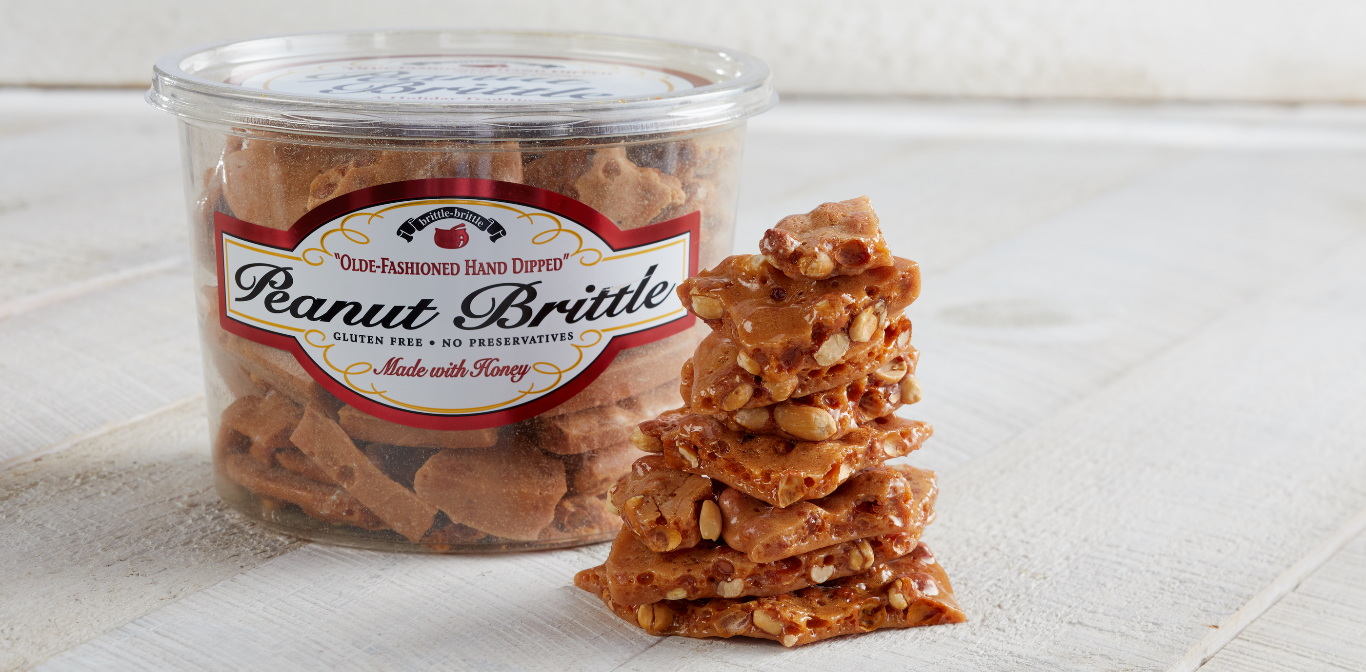 A clear plastic tub of Old Fashioned Hand Dipped Peanut Brittle next to a tall stack of glazed peanut brittle pieces on a white wooden surface.