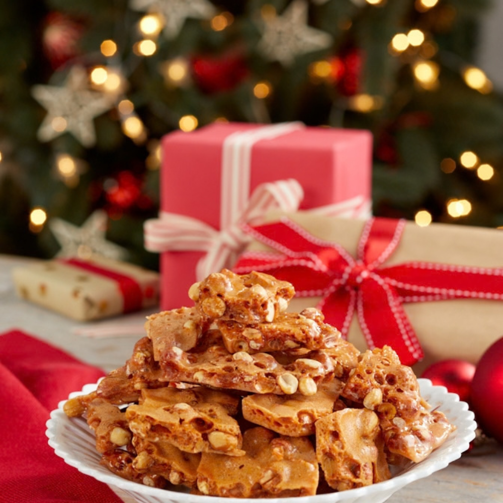 Brittle-Brittle peanut brittle piled in a white dish in front of wrapped holiday gifts and a lit Christmas tree