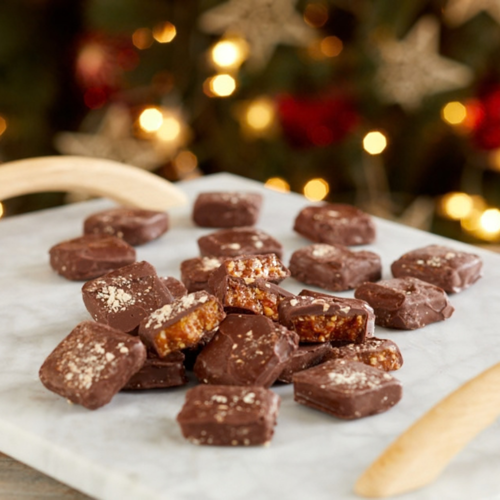 Brittle-Brittle Roasted Almond Toffee pieces covered in dark chocolate on a marble board, holiday lights behind